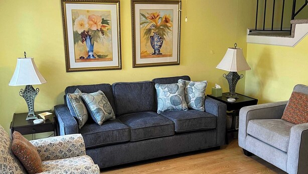 Una sala de estar con un sofá gris, sillones estampados, una mesa de comedor de madera y una escalera.
