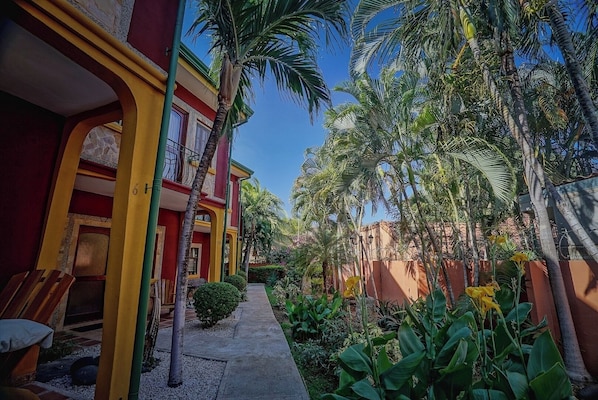 A multi-story building with a red and yellow facade, a narrow pathway, and lush greenery including palm trees and tropical plants.
