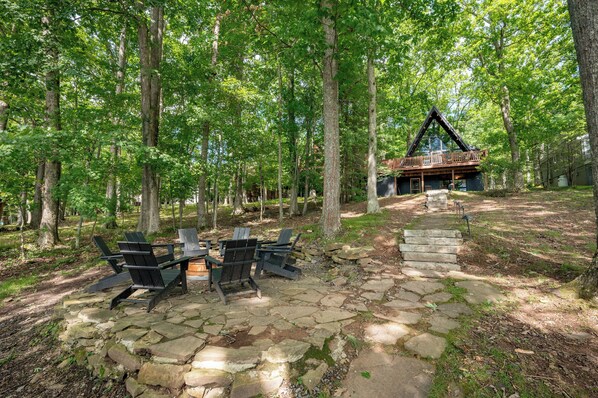 Une cabane dotée d’une terrasse en pierre et de fauteuils Adirondack, entourée d’une forêt.