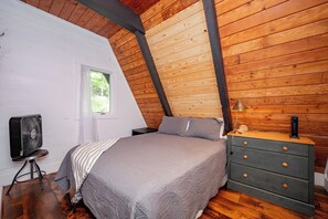 A bedroom with a sloped ceiling, a bed, a dresser, a fan, and a window.