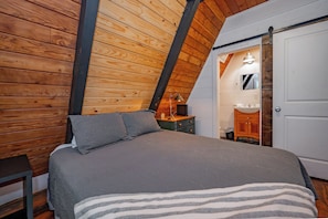 A bedroom with a sloped ceiling, wooden walls, a bed with grey bedding, a nightstand, a lamp, and a bathroom with a mirror and sink.