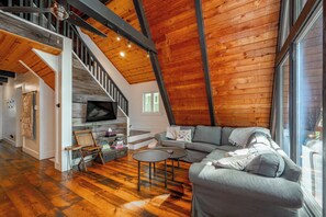 A spacious loft with a high ceiling, wooden beams, a staircase, a living area with a sofa and coffee table, and a television mounted on the wall.