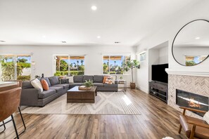 A modern living room with a sectional sofa, a coffee table, a fireplace, and large windows offering a view of palm trees.