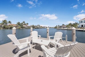 A deck with white Adirondack chairs and a table, overlooking a body of water with palm trees and buildings in the background.
