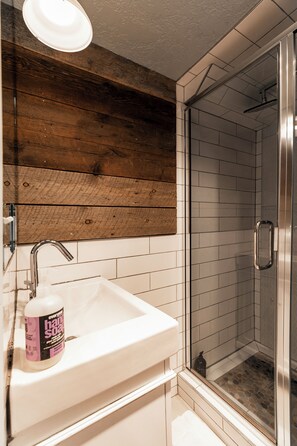 A bathroom with a white sink, wooden wall paneling, a glass shower enclosure, and a bottle of hand soap. - Acorn Chalet Studio | 100 Yards to Park City Mountain | Sleeps 2 (Park City)