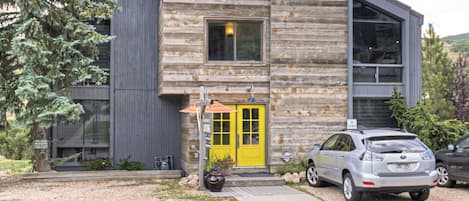 Uma cabana de toras moderna com porta amarela, uma janela grande e um carro estacionado na frente.
