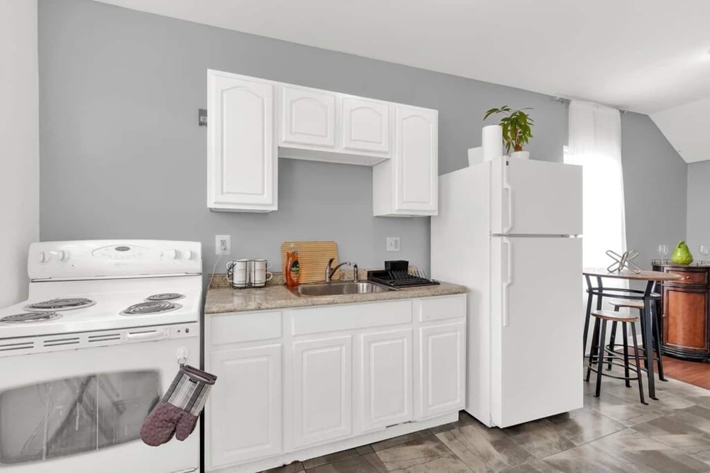 A modern kitchen with white cabinetry, a stove, a sink, and a refrigerator. There is a wooden cutting board and a potted plant on the counter.