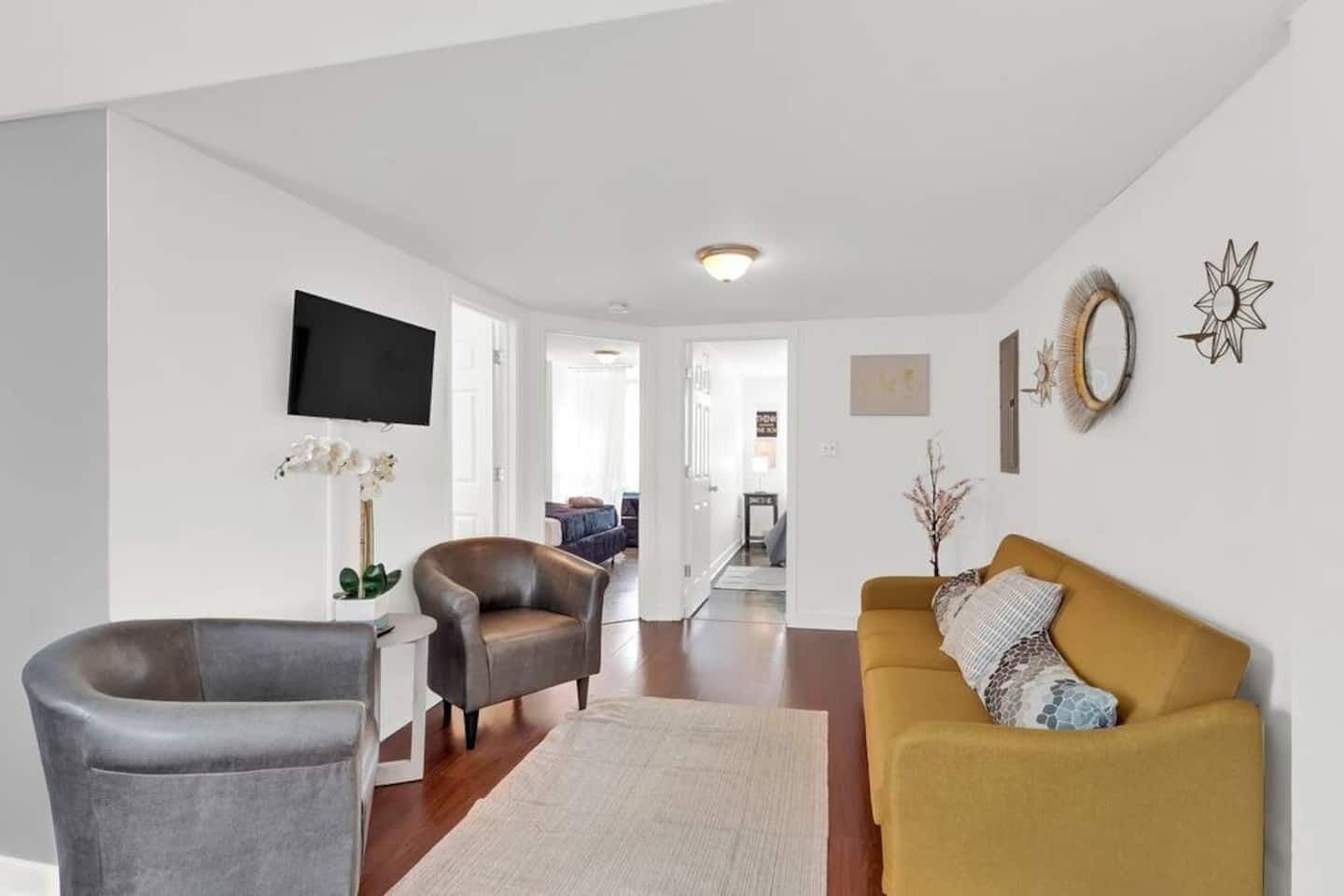 A modern living room with a grey armchair, a brown leather chair, and a mustard-colored sofa. There is a flat-screen TV mounted on the wall, a small table with a vase of flowers, and a decorative mirror.