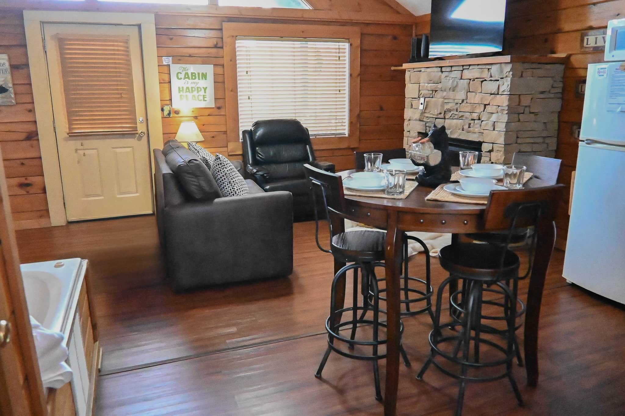 L’intérieur chaleureux d’un chalet comprend une table à manger pour quatre personnes, un canapé en cuir noir, une cheminée en pierre et un téléviseur fixé au mur.