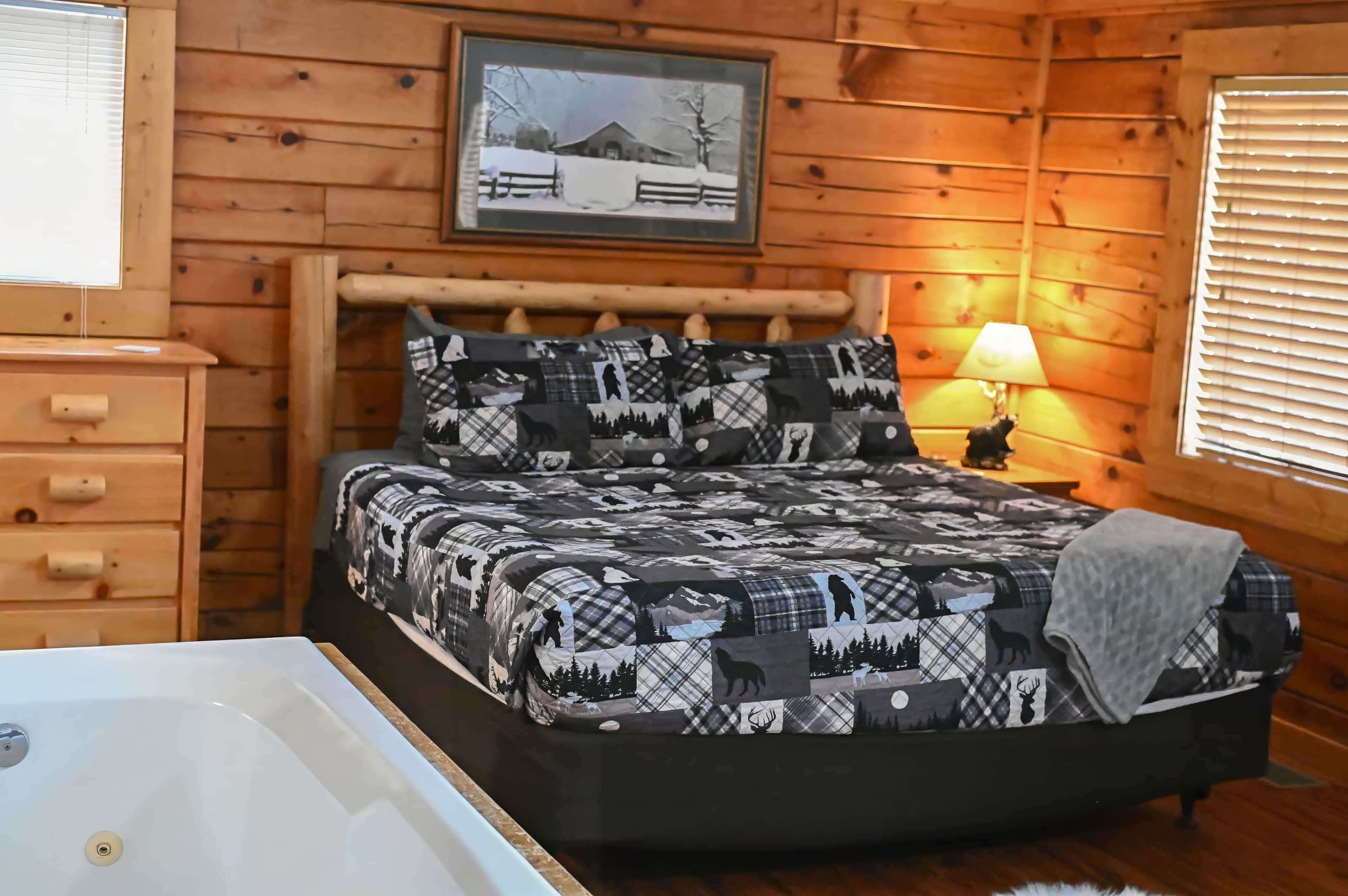 A cozy cabin room with a bed featuring a forest and wildlife print, a wooden dresser, a window with blinds, and a framed picture on the wall.