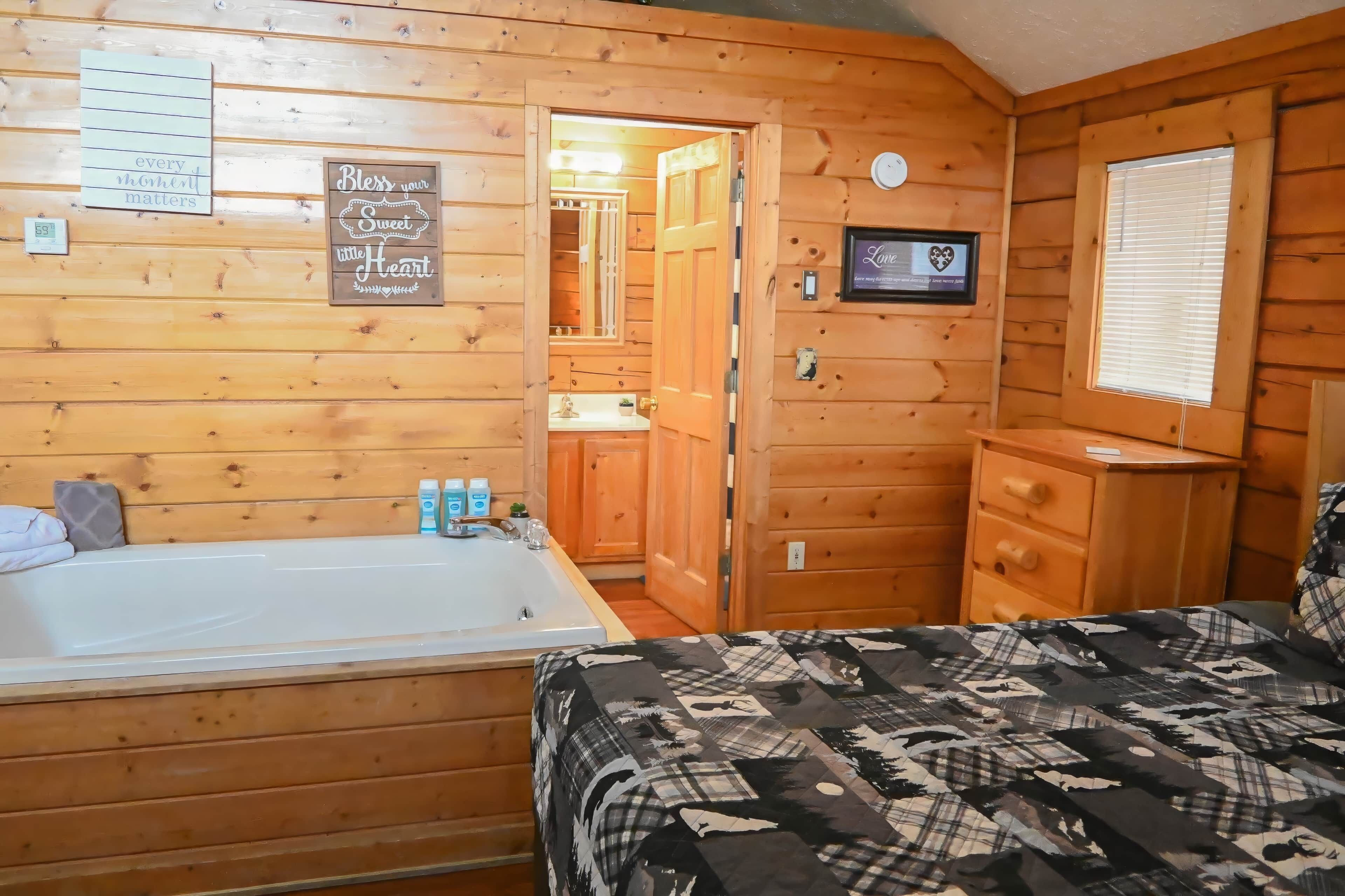 Une chambre de style cabane en bois, avec une grande baignoire, un lit recouvert d’une couverture à motifs, une commode en bois et une petite fenêtre avec des stores.