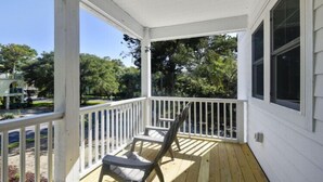 A covered porch with a wooden deck, a single chair, and a view of a green landscape. - Updated and Modern Corolla Beach Retreat! (Corolla)