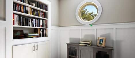A room with a built-in bookshelf, a small cabinet with glass doors, and a round window with a view of trees.