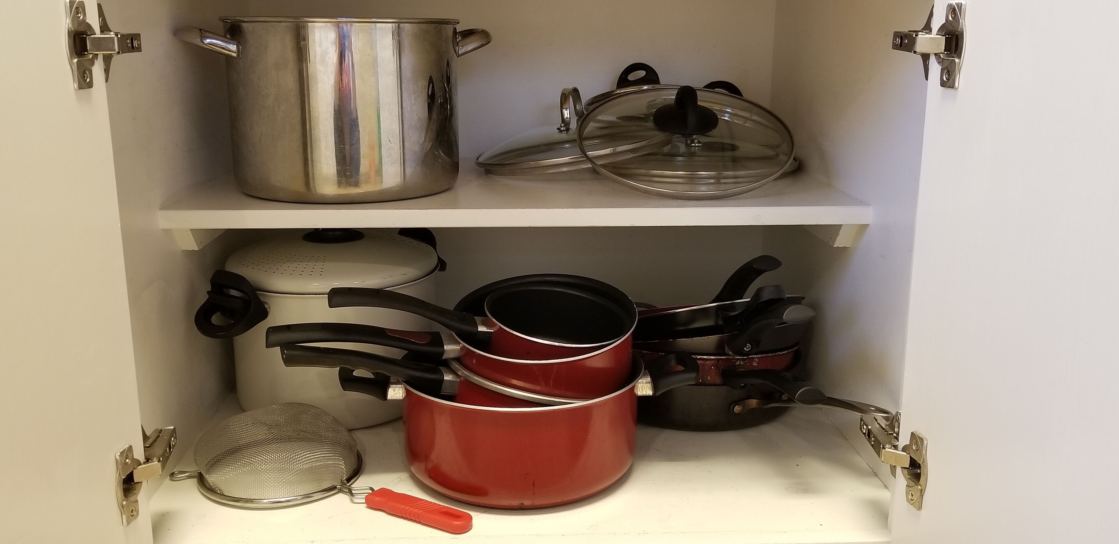 A kitchen cabinet with stainless steel pots, red and black pots, and a strainer.