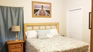 A bedroom with a wooden headboard, a bed with floral bedding, a nightstand with a lamp, a framed picture on the wall, and a white door.