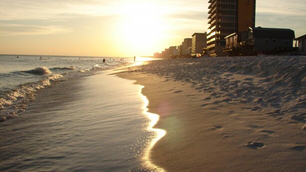 A beach with a sunset, waves, and buildings in the background.