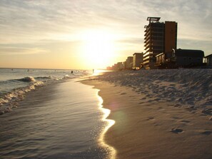 A beach with a sunset, waves, and buildings in the background.