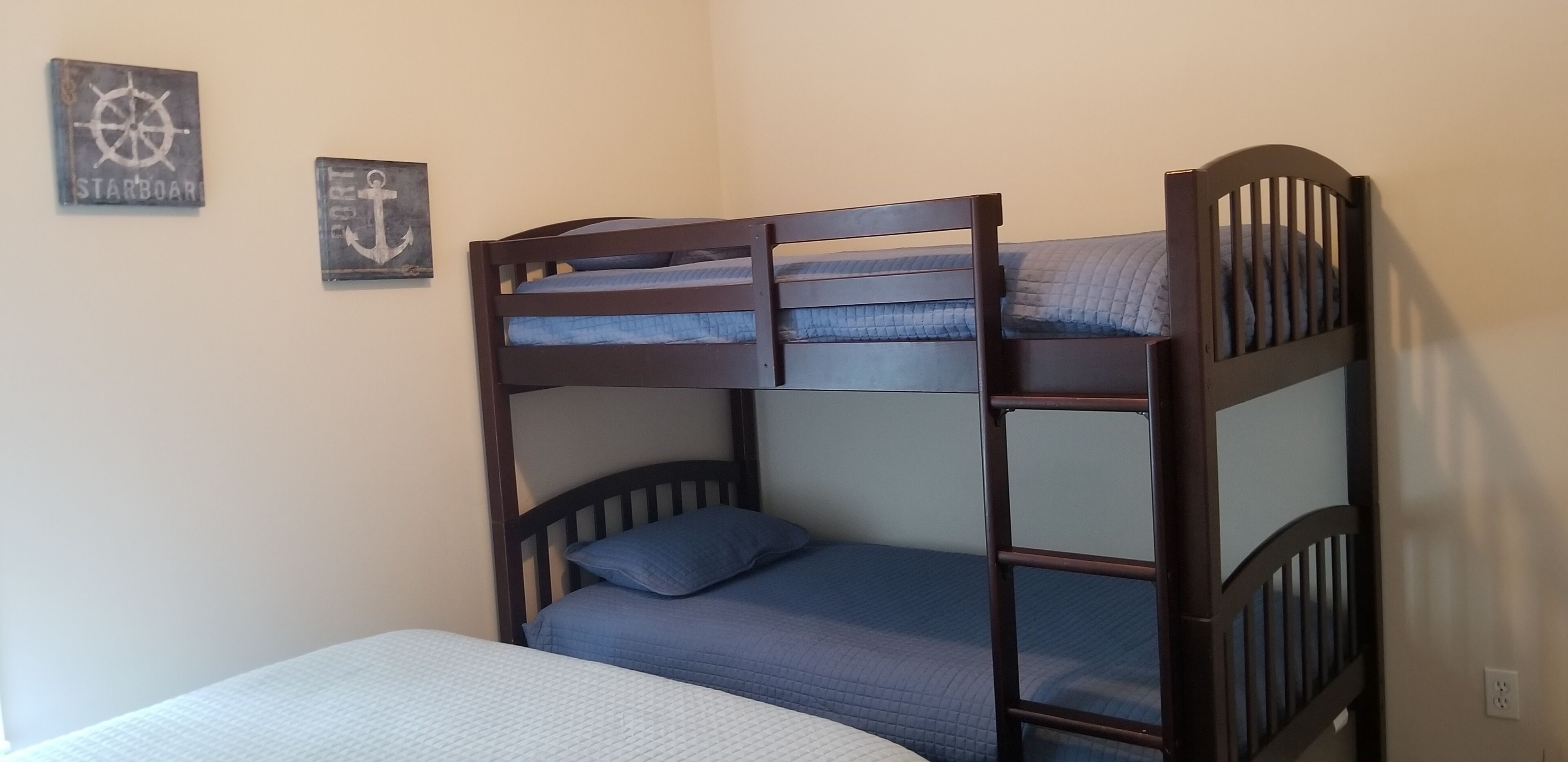 A bunk bed with a lower bunk and a single bed, both with blue bedding. There are two wall decorations: one with a ship's wheel and the other with an anchor.