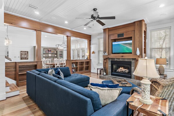 A modern living room with a sectional sofa, a fireplace, a television, and a dining area with a view of the outdoors. - Park Front-Camp Watercolor! 6 bikes 4 Bedrooms (Santa Rosa Beach)