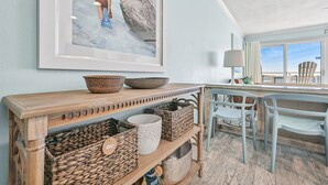 Ein Zimmer mit einem Holzkonsolentisch, der gewebte Körbe und eine Schale beherbergt, einem gerahmten Bild von zwei Personen auf einem Felsen, einer Lampe und Meerblick durch ein Fenster.