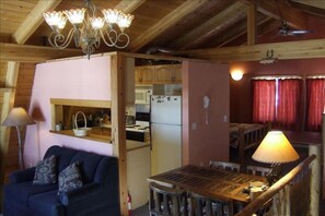 A cozy cabin interior with a wooden ceiling, a kitchenette, a dining area with a table and chairs, and a sofa with patterned cushions.