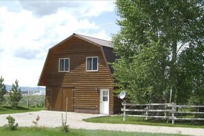 A wooden barn with a large door and two windows, surrounded by a gravel driveway and a wooden fence.