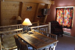 A rustic wooden room with a table and chairs, a lamp, a television, and a colorful curtain.