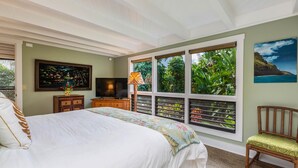 A bedroom with a bed, a TV, a wooden dresser, and a window with a view of greenery.
