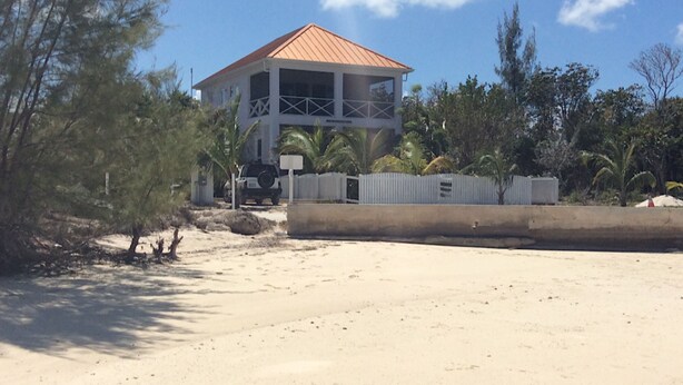 A two-story building with a red roof, surrounded by trees and a sandy area.