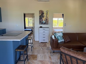 A modern interior with a kitchen island, a dining area, a living room with a brown leather sofa, and a large window with a view of greenery.