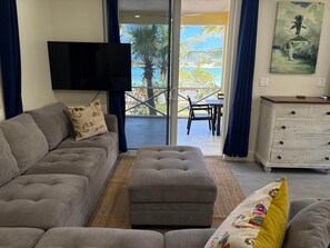 A living room with a grey sofa, a matching ottoman, a flat-screen TV mounted on the wall, a wooden cabinet with drawers, and a view of a balcony with outdoor furniture.