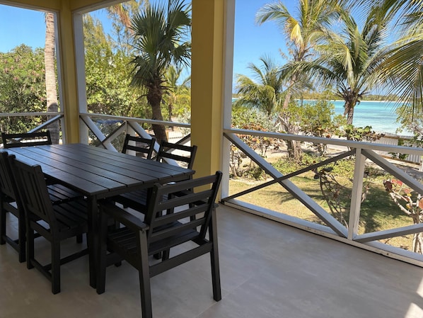 A covered outdoor dining area with a table and chairs, a view of palm trees and a beach.