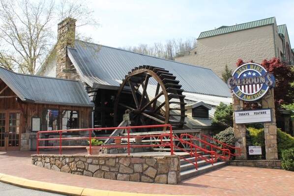 Una histórica rueda hidráulica con base de piedra y barandillas rojas, un edificio con techo de metal y un letrero que indica un restaurante llamado 'Calhoun's'.