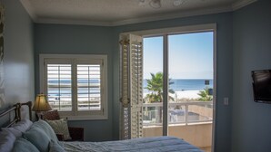 Quarto com uma cama grande, janela com persianas, varanda com vista para a praia e televisão na parede.