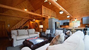 L’intérieur chaleureux d’un chalet présente un salon avec un canapé, des fauteuils et une cheminée. On aperçoit à l’arrière-plan un coin cuisine équipé d’appareils électroménagers modernes.