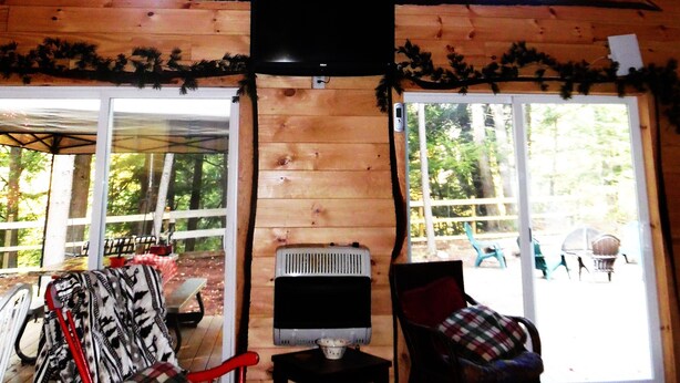A cabin with a sloped wooden ceiling, a television mounted on the wall, and two windows providing natural light. - "Your Neck of the Woods" Cabin Rental: Peaceful Get-away Overlooking Fish Creek (Taberg)