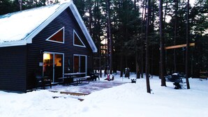A cabin with a snow-covered roof, a porch with benches, and a forested backdrop. - "Your Neck of the Woods" Cabin Rental: Peaceful Get-away Overlooking Fish Creek (Taberg)