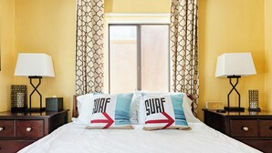 Habitación de hotel con una cama, dos mesitas de noche, dos lámparas, una ventana con cortinas y dos almohadas con la palabra 'SURF'.