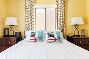 Una camera d'albergo con un letto, due comodini, due lampade, una finestra con tende e due cuscini con la scritta 'SURF'.