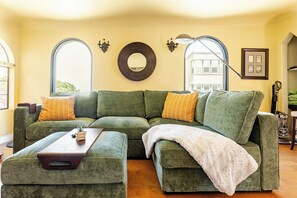 A green sectional sofa with a white throw blanket and two side tables, one with a plant and the other with a remote control.