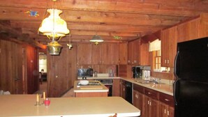 Interior de uma cabana de madeira com cozinha, incluindo armários de madeira, geladeira, micro-ondas e mesa de jantar com duas cadeiras.