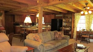 Uma sala de estar acolhedora com sofá, poltrona e mesa de centro. Uma cozinha é visível ao fundo, com geladeira, fogão e mesa de jantar.
