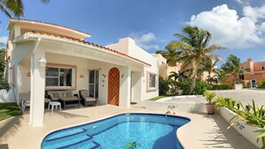 Ein Pool mit gewölbter Kante, eine überdachte Terrasse mit Gartenmöbeln und eine Palme im Hintergrund.