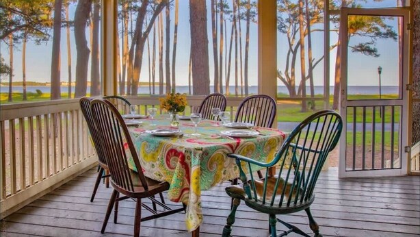 A covered outdoor dining area with a table set for four, surrounded by chairs, overlooking a body of water and trees.