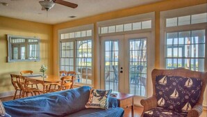 A living room with a ceiling fan, a blue sofa, a patterned armchair, a dining table with chairs, and large windows with a view of the outdoors.