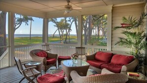 A covered outdoor seating area with wicker furniture and red cushions, overlooking a grassy landscape and trees.