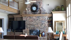 A living room with a stone fireplace, a ceiling fan, a flat-screen TV, and a dining area with a table and chairs. - Gorgeous Home In Upscale Neighborhood - Room For The Whole Family 4 bedrooms! (Pawhuska)