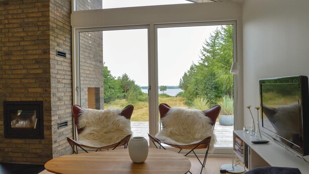 A modern living room with a large sliding glass door leading to a view of a lake and trees. There are two white furry chairs, a wooden table, and a flat-screen TV on a stand.