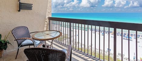 A balcony with a glass table and wicker chairs, overlooking a beach with umbrellas and people.