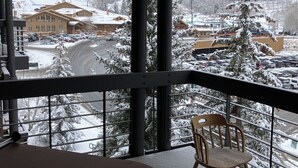 A snow-covered outdoor area with a wooden chair and a table, overlooking a parking lot and buildings. - Deer Valley-100yds~1min walk. Closest Powder Run Penthouse. Deck Hot Tub w'Views (Park City)